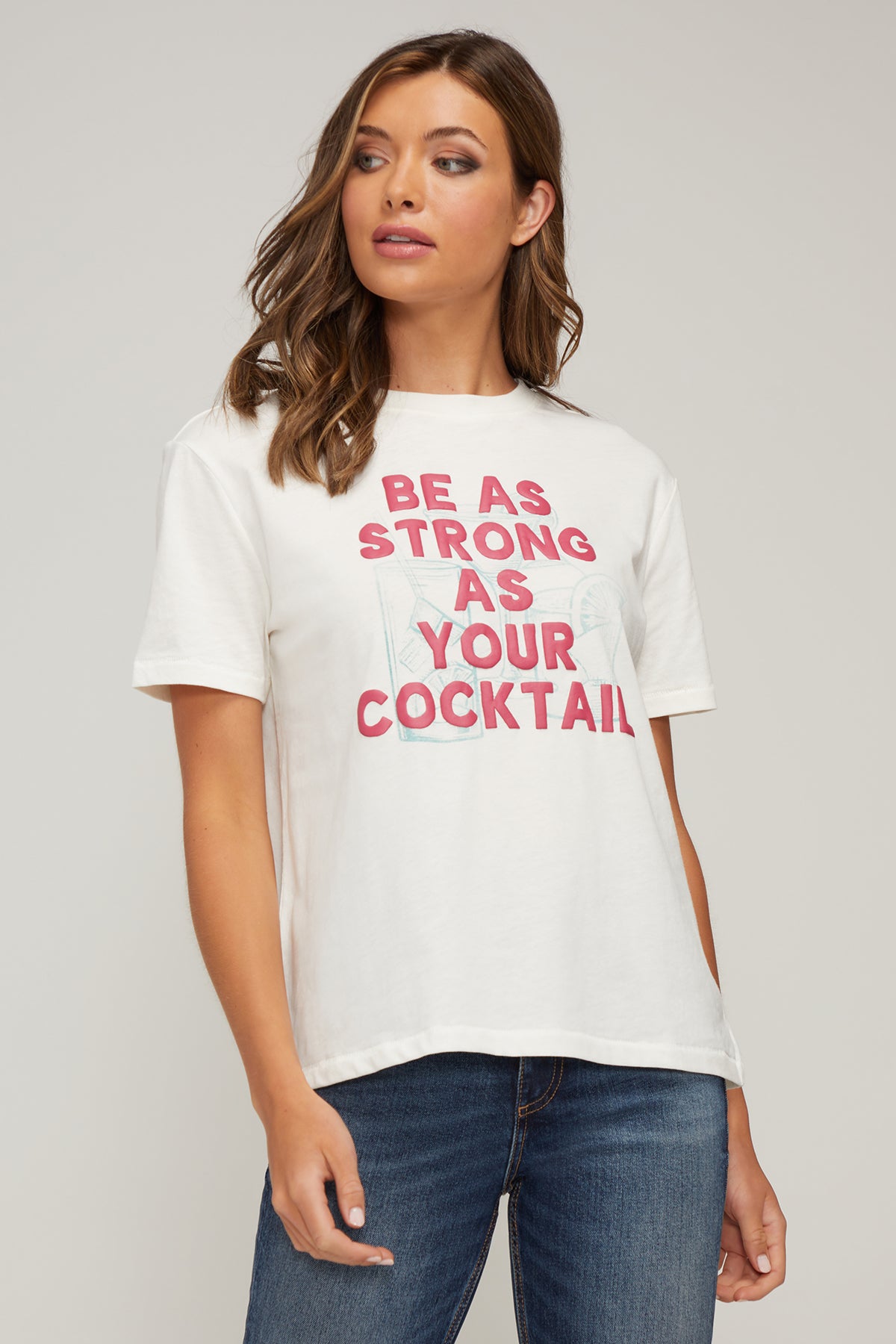 A woman stands against a neutral gray background, wearing a casual outfit. She has medium-length brown hair and wears a white T-shirt with bold red text that reads &quot;BE AS STRONG AS YOUR COCKTAIL.&quot; Below the text is a faint outline of a cocktail glass with a lemon slice. She pairs the T-shirt with blue denim jeans, standing with one hand relaxed at her side and the other slightly raised. The overall look is casual and confident, perfect for a relaxed, stylish appearance.